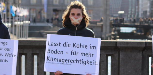Eine Person vor einem Geländer mit einem großen Schild in den Händen. Auf dem Schild steht: