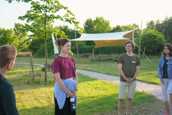 Junge Menschen stehen drau&szlig;en, im Hintergrund ein Weg und ein Pavillion, eine junge Person spricht l&auml;chelnd