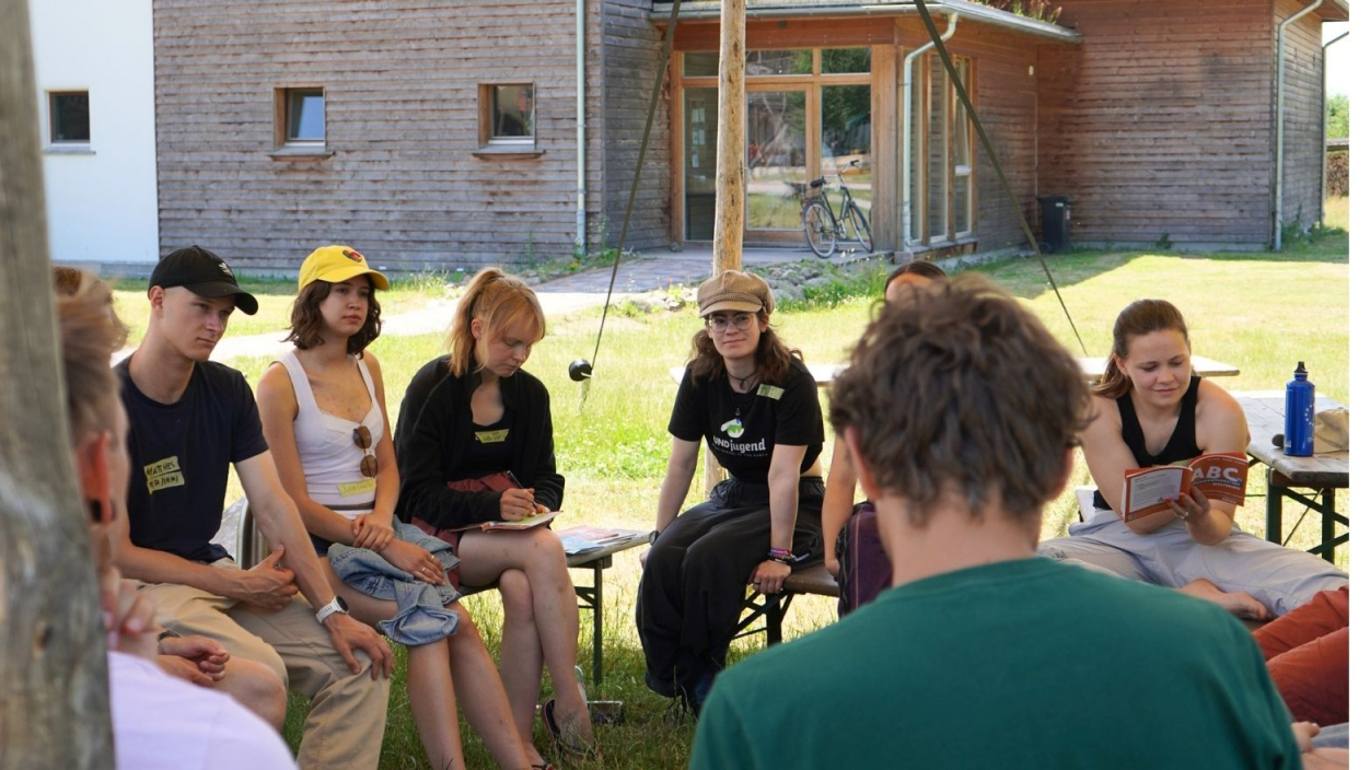 Eine Gruppe junger Menschen sitzt im Sommer unter einem Baldachin auf Holzbänken und tauscht sich aus