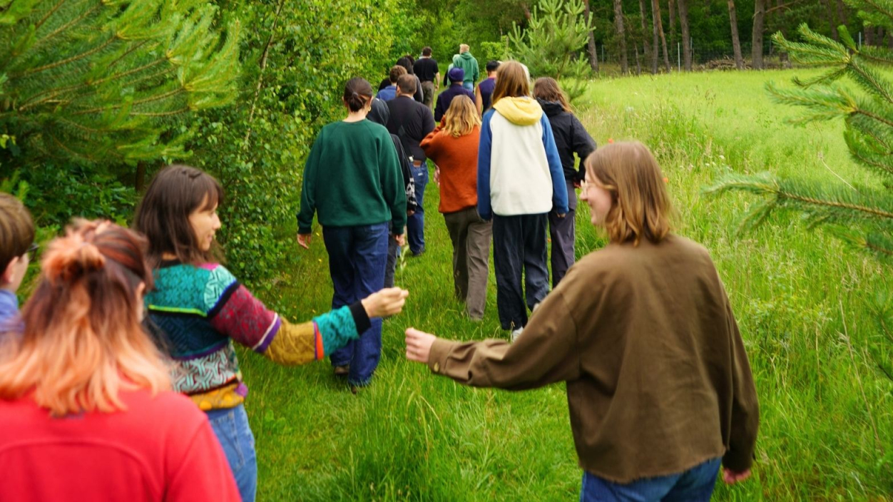Eine Gruppe geht zwischen Wald und Wiese spazieren, zwei junge Personen geben sich im Laufen schlagen sich lachend in die Hände
